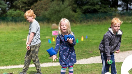 A young girl and two young boys taking part in a bean bag throwing competition outdoors in the sunshine
